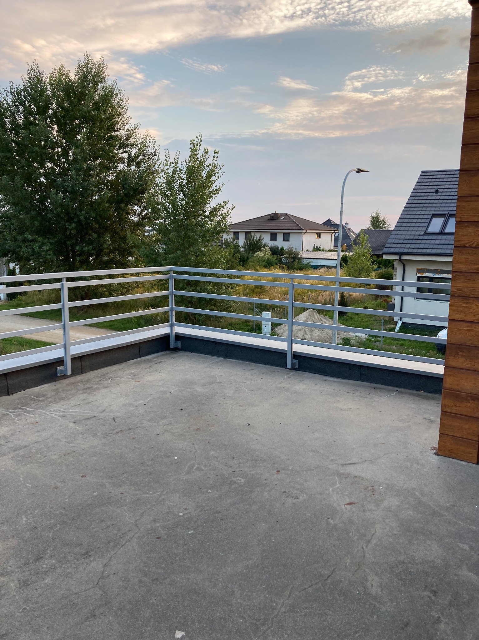 Szara, metalowa balustrada balkonowa z trzema poprzeczkami na tle domów jednorodzinnych i zachmurzonego nieba o zmierzchu.