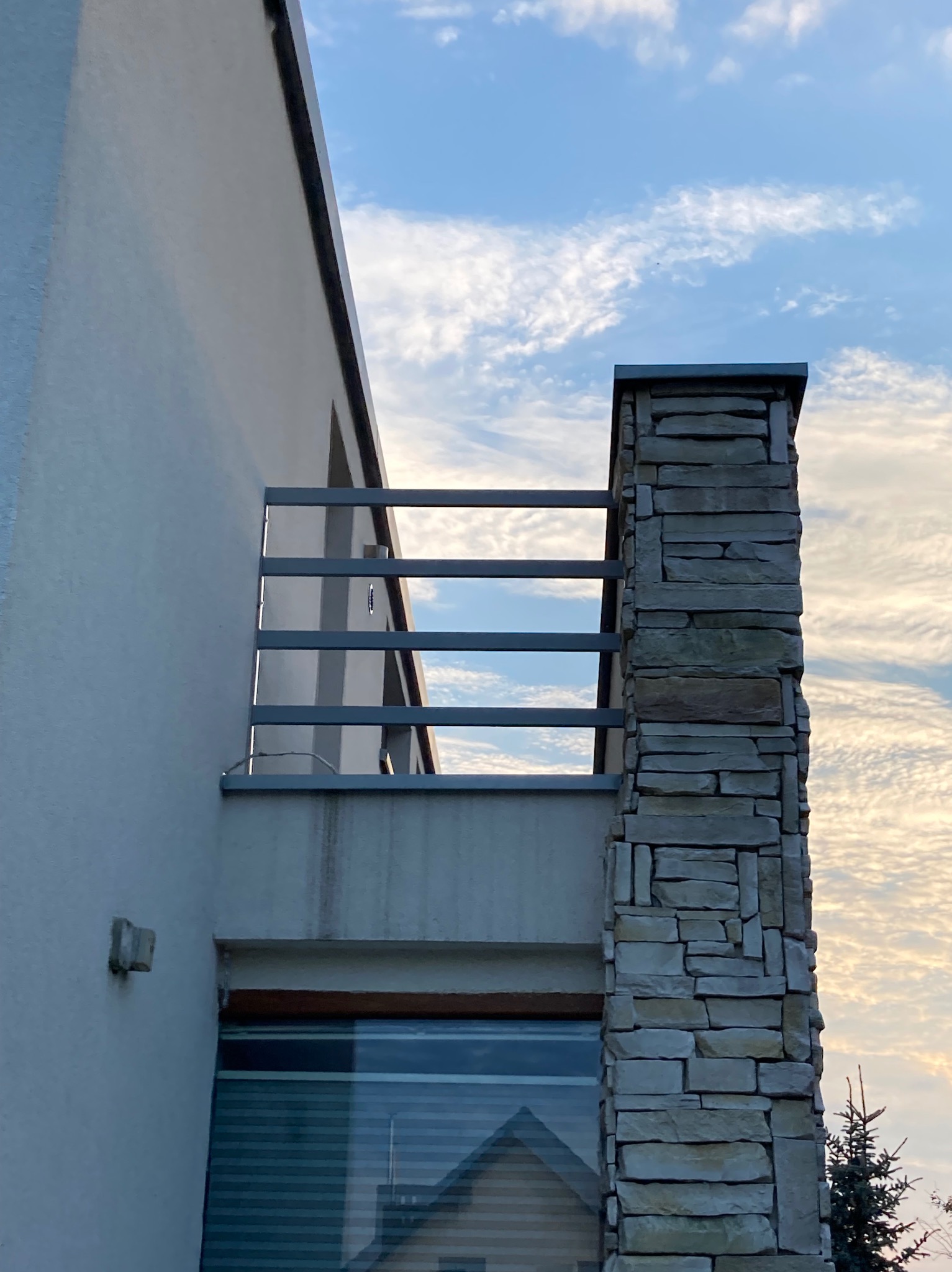 Nowoczesny balkon z prostą, metalową barierką i kamiennym filarem na tle błękitnego nieba z chmurami.