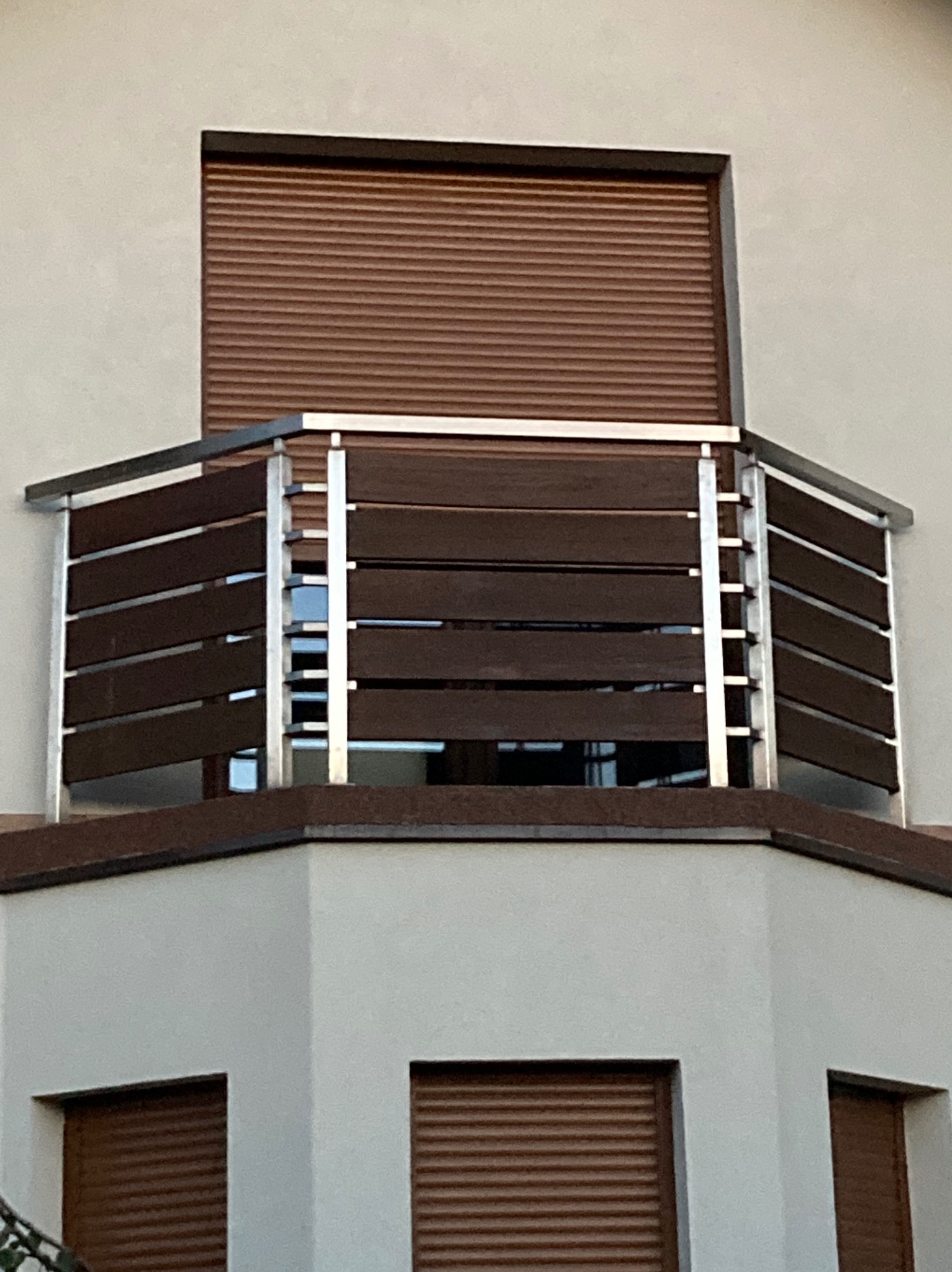 Nowoczesna balustrada balkonowa z poziomych, ciemnobrązowych paneli drewnopodobnych i srebrnych, metalowych słupków, z widocznymi roletami okiennymi w tle.