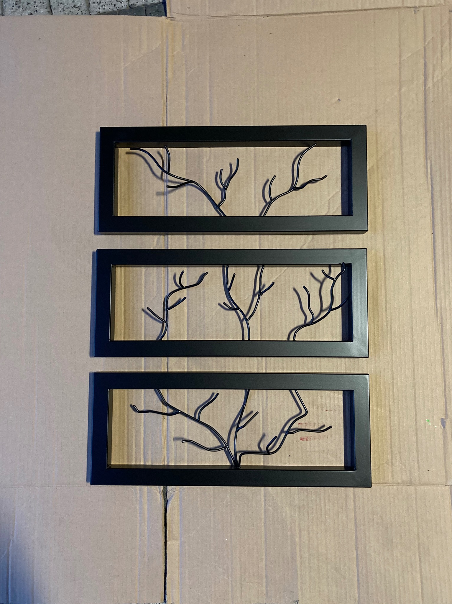 Trzy czarne, prostokątne ramy z metalowymi, stylizowanymi drzewami w środku, ułożone pionowo na kartonowym tle.