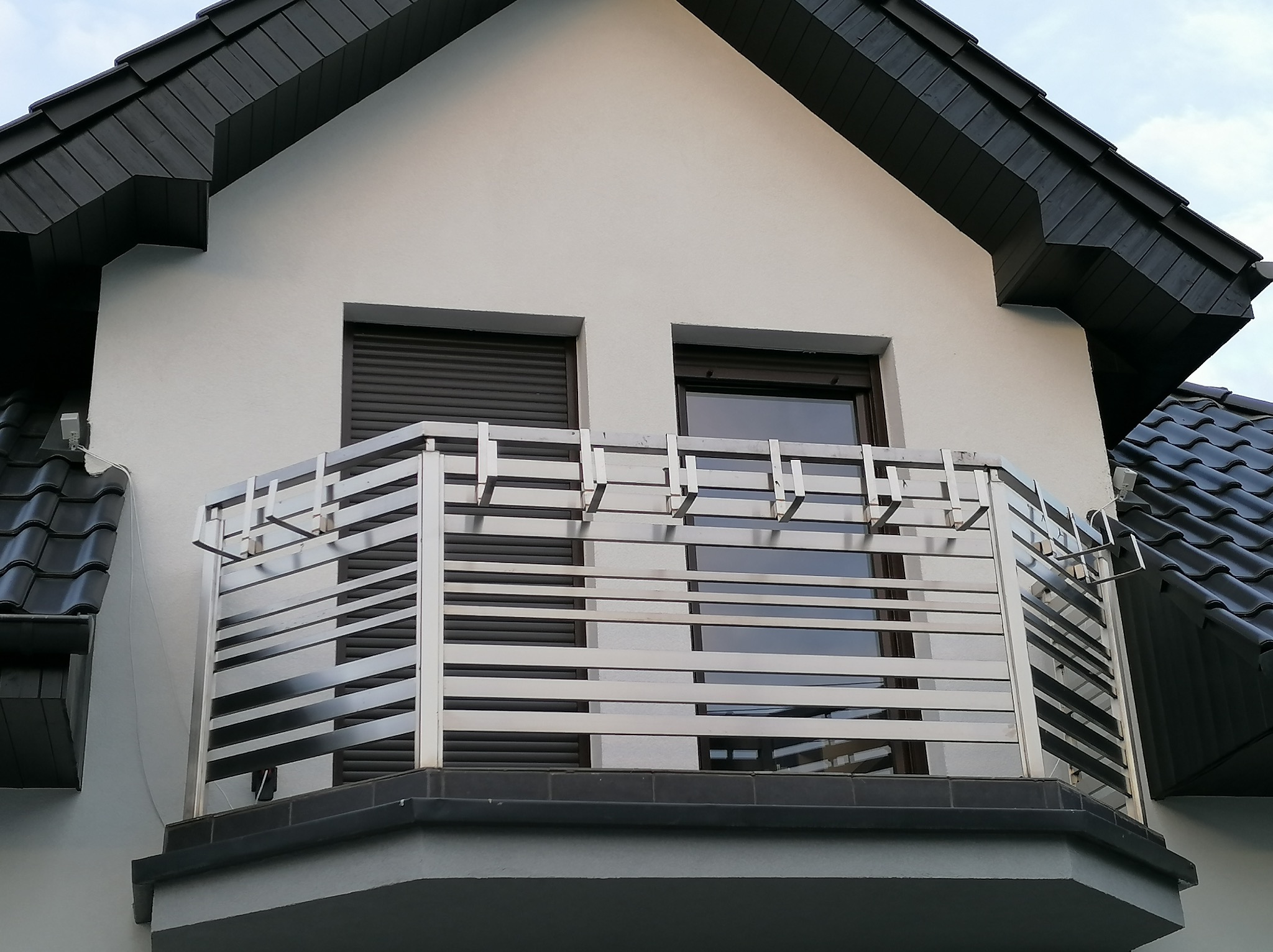 Nowoczesna balustrada balkonowa ze stali nierdzewnej z poziomymi, prostokątnymi elementami, zamontowana na balkonie domu z ciemną dachówką i szarymi roletami okiennymi.