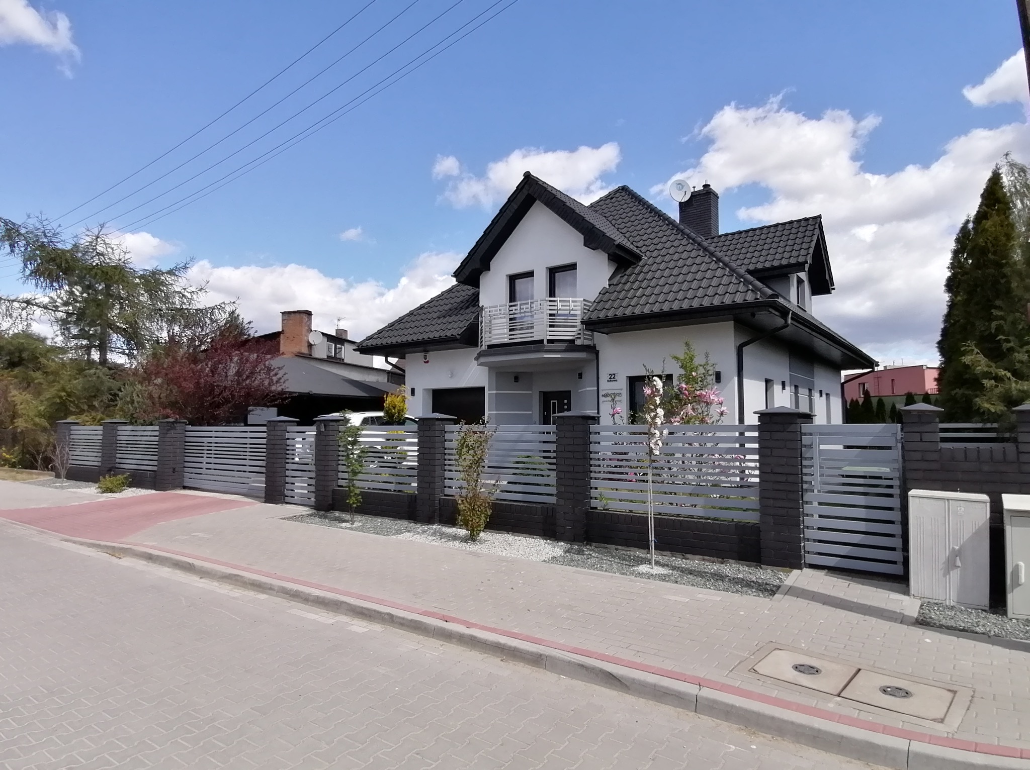 Nowoczesny dom jednorodzinny z szarą, poziomą bramą i ogrodzeniem z elementami z cegły klinkierowej, na tle błękitnego nieba z chmurami.