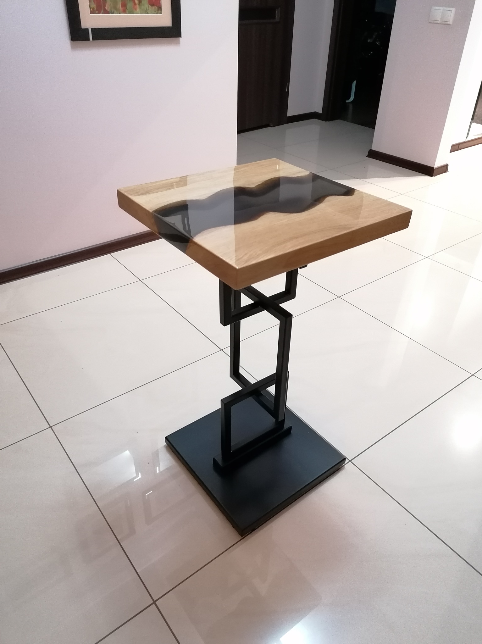 Stolik kawowy z blatem z drewna i żywicy epoksydowej, na metalowej podstawie o geometrycznym kształcie, ustawiony na jasnych płytkach podłogowych.