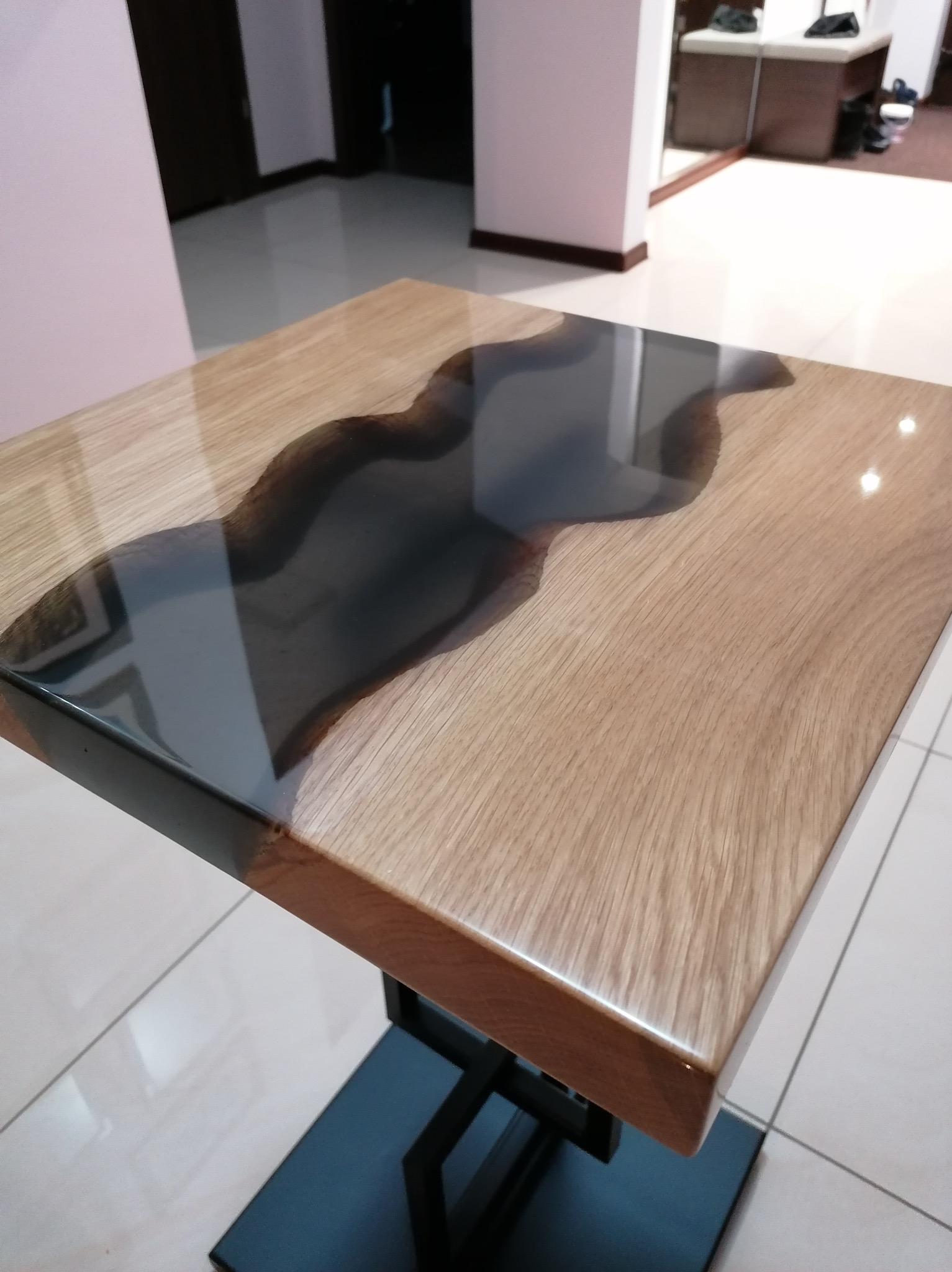 Kwadratowy stolik kawowy z blatem z jasnego drewna dębowego i wtopioną, nieregularną żywicą epoksydową w kolorze antracytowym, osadzony na metalowej, czarnej podstawie w kształcie litery X.