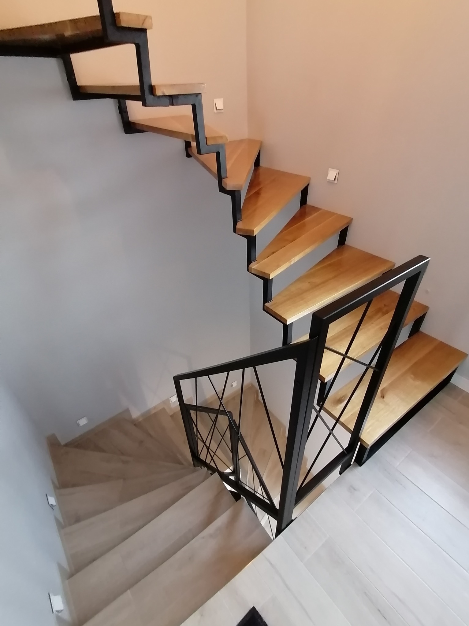 Nowoczesne schody z drewnianymi stopniami i czarną, metalową balustradą o geometrycznym wzorze, zamontowane w jasnym wnętrzu z szarymi ścianami i drewnopodobną podłogą.