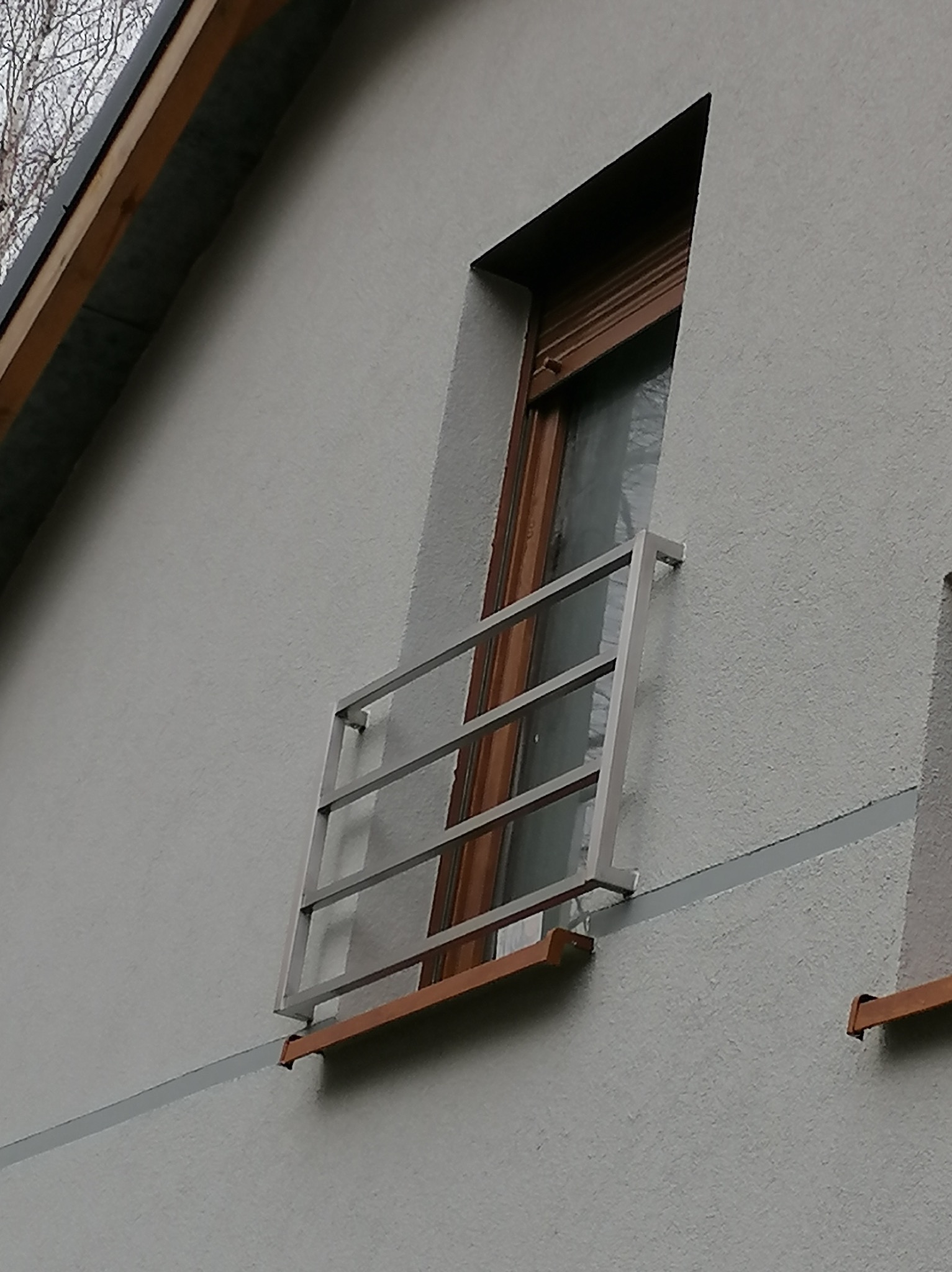 Metalowa balustrada okienna z trzema poprzeczkami zamontowana na szarej elewacji budynku z widocznym oknem z brązową ramą.