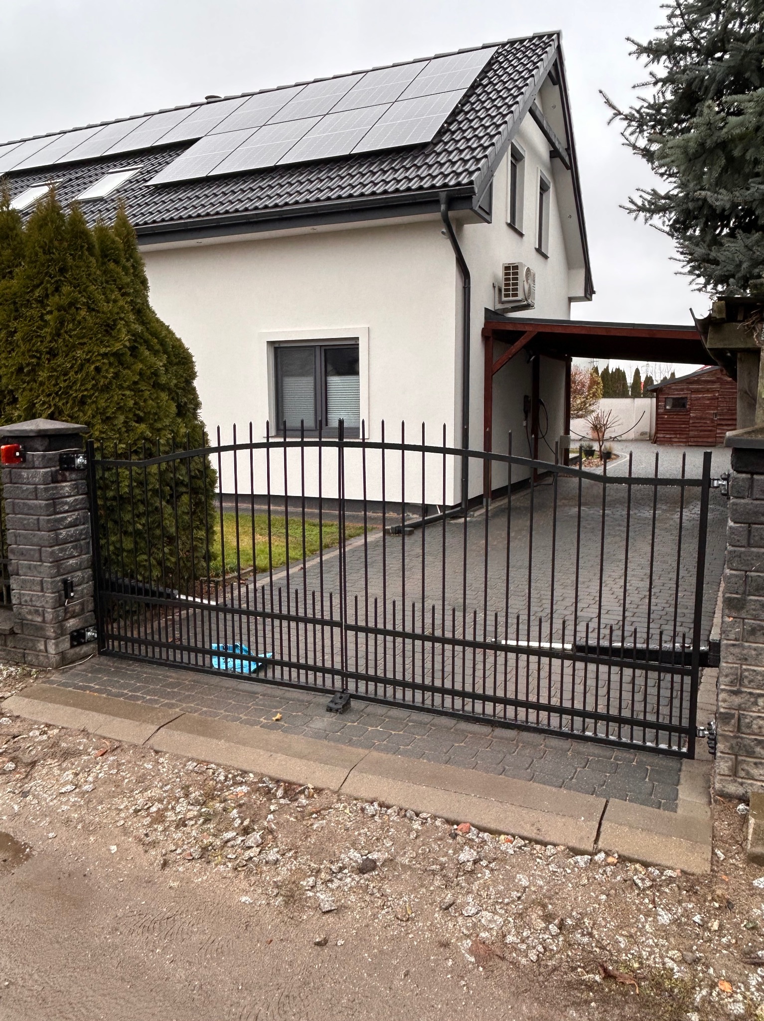 Czarna brama wjazdowa z pionowymi prętami, zamontowana na ceglanych słupkach, na tle domu z panelami słonecznymi na dachu.