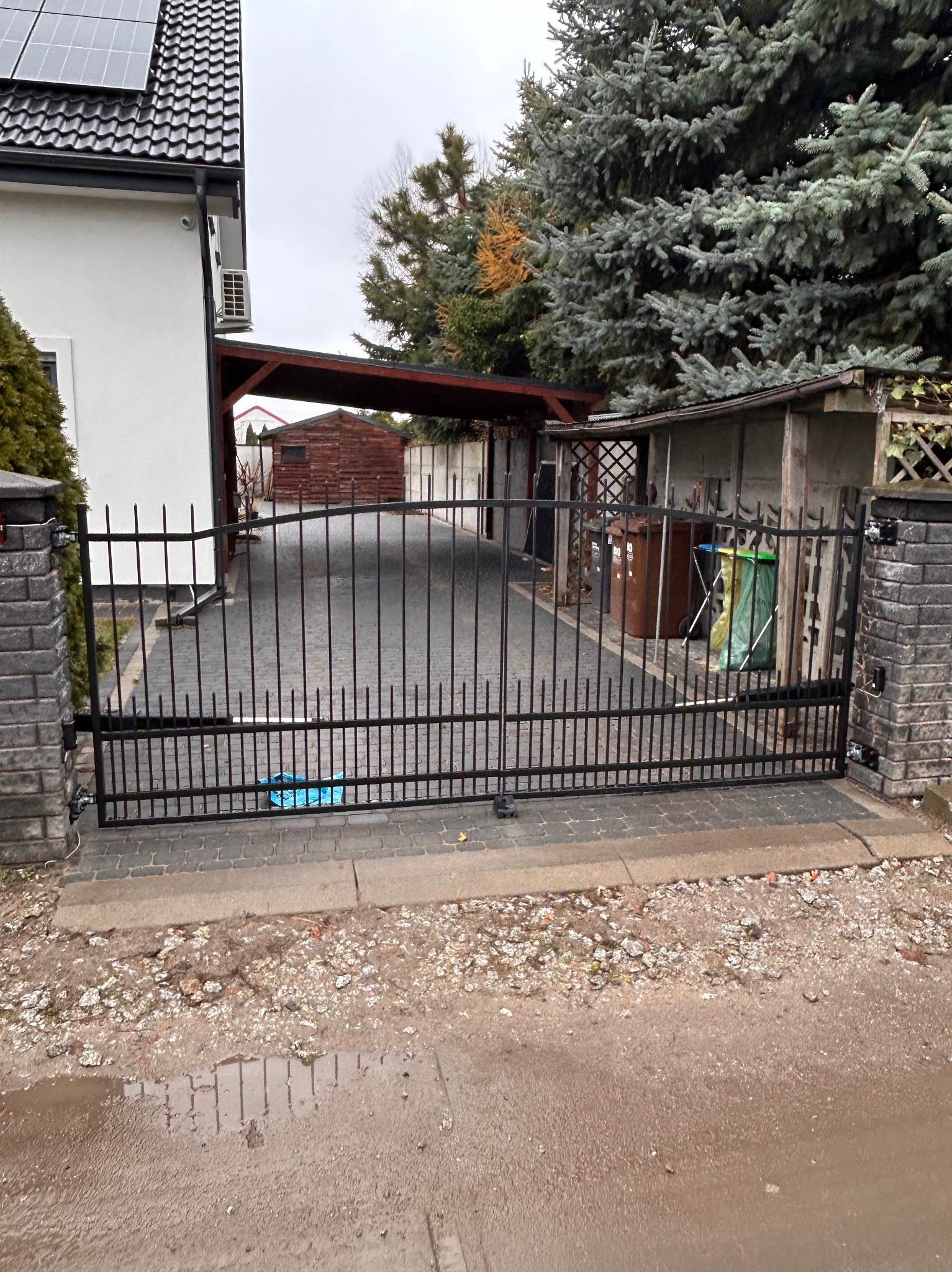 Czarna metalowa brama wjazdowa z pionowymi prętami, zamontowana między murowanymi słupkami, z widokiem na podjazd z kostki brukowej i budynek mieszkalny z panelami słonecznymi na dachu w tle,...