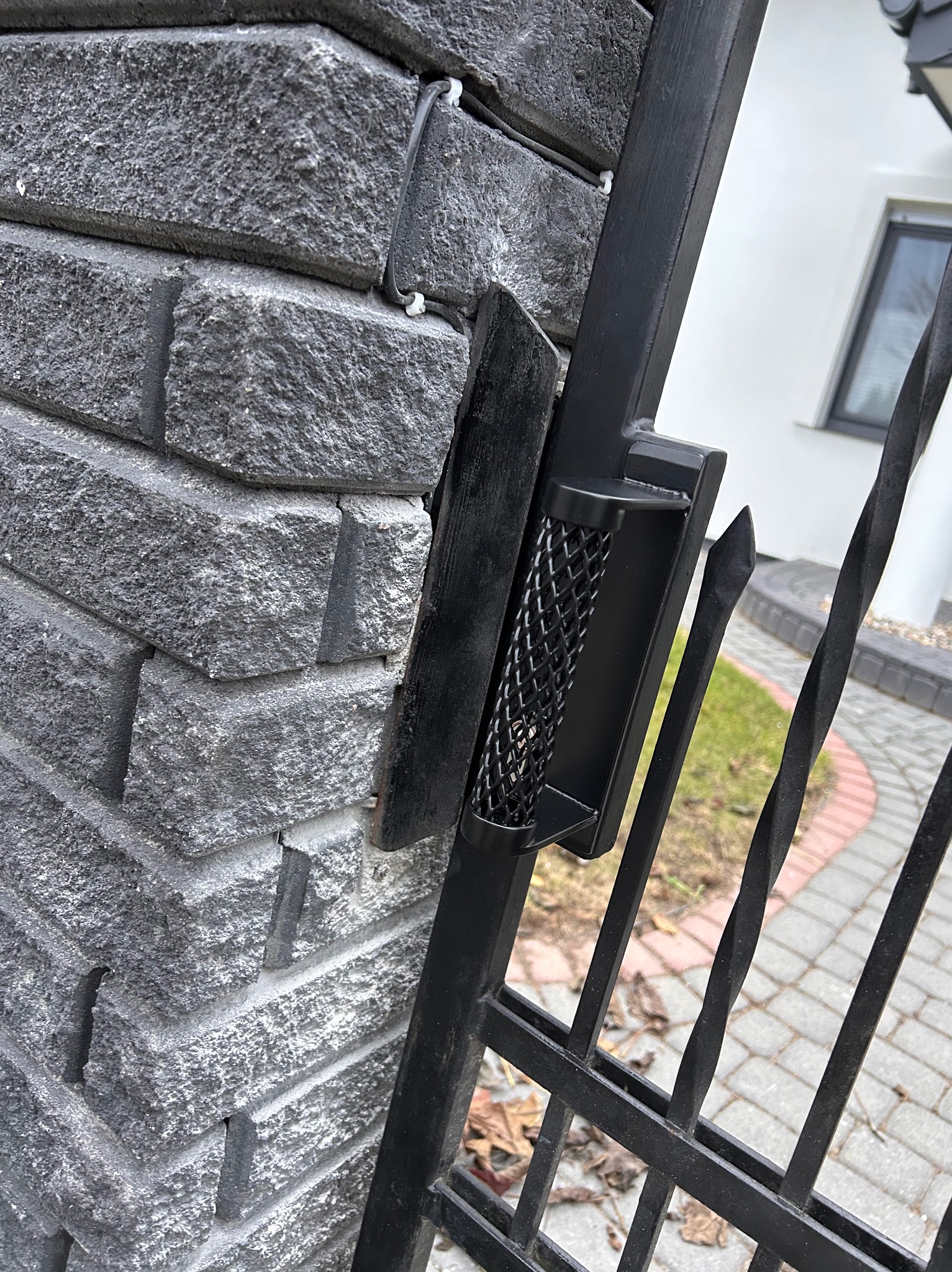 Czarny, metalowy uchwyt z siatkową fakturą na skrzydle bramy, zamontowany do szarego, kamiennego słupka ogrodzeniowego z widocznym fragmentem posesji w tle.
