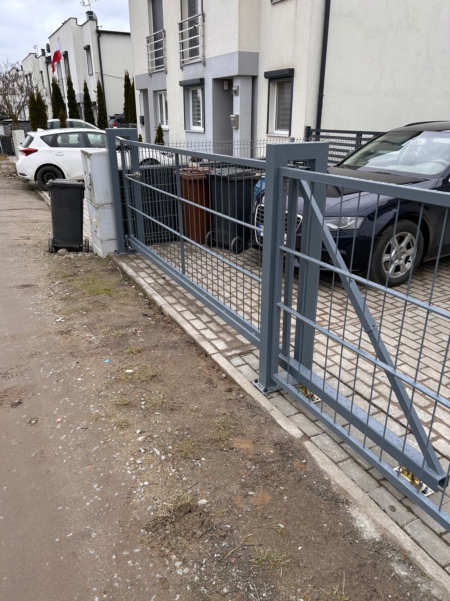 Szara, przesuwna brama wjazdowa z metalową siatką na posesji z kostki brukowej, w tle zaparkowane samochody i szereg domów.