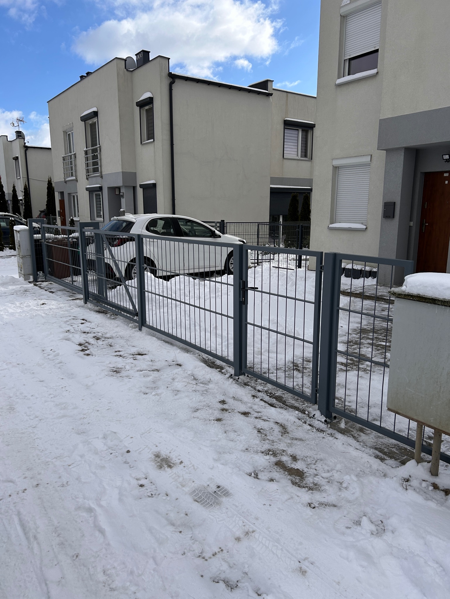 Szara, metalowa brama wjazdowa z pionowymi prętami, z widocznym fragmentem białego samochodu i zasypanym śniegiem podjazdem na tle nowoczesnych domów.