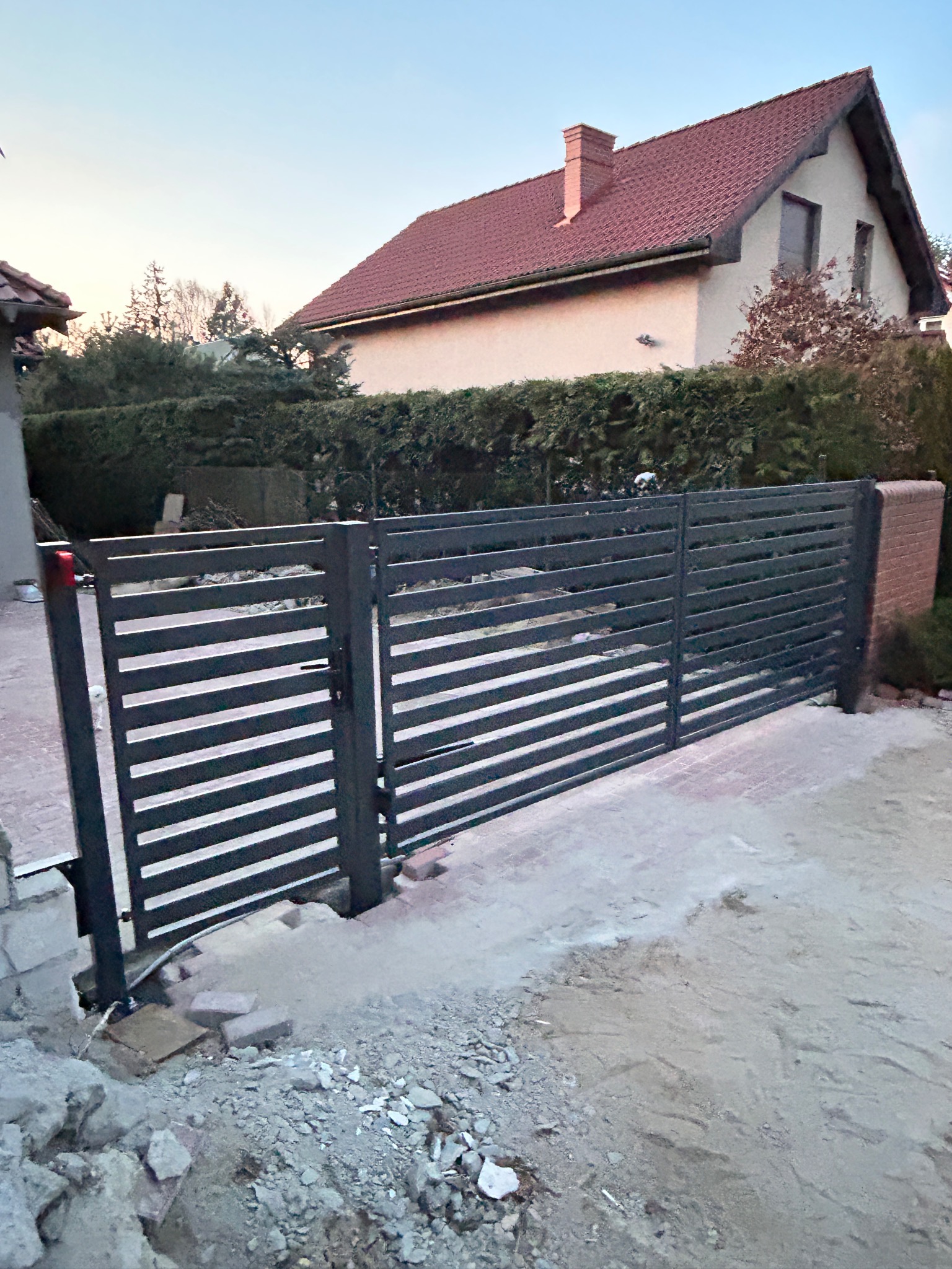 Nowoczesna brama wjazdowa przesuwna z poziomych profili, w trakcie montażu, widoczne niedokończone podłoże i fragment domu w tle.
