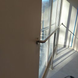 RAF-SPAW - Wnętrze budynku z przeszkloną ścianą i zamontowaną metalową poręczą, widoczne światło słoneczne padające na podłogę i ścianę, kabel elektryczny leżący na podłodze.