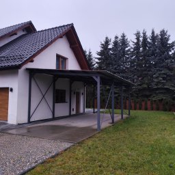 RAF-SPAW - Nowoczesna, metalowa pergola przy białym domu z drewnianą bramą garażową. Szara konstrukcja z zadaszeniem, widoczne elementy mocujące. Tło: zielony trawnik i wysokie drzewa.