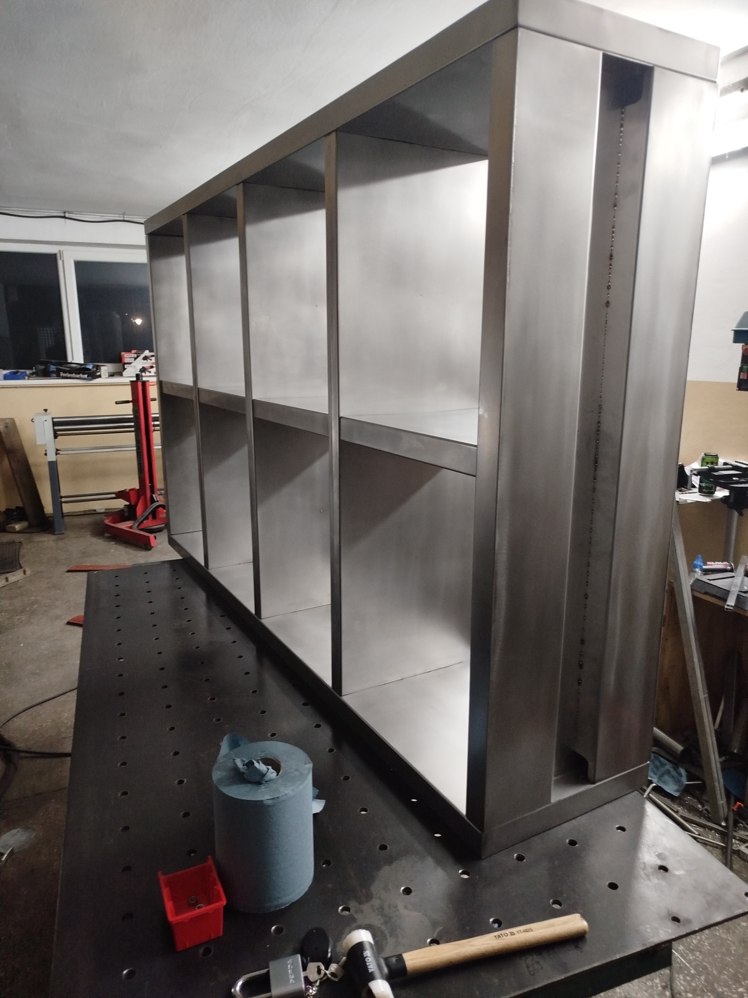 Surowa, metalowa konstrukcja regału z półkami, widoczna w warsztacie na stole spawalniczym, obok narzędzia i akcesoria montażowe.