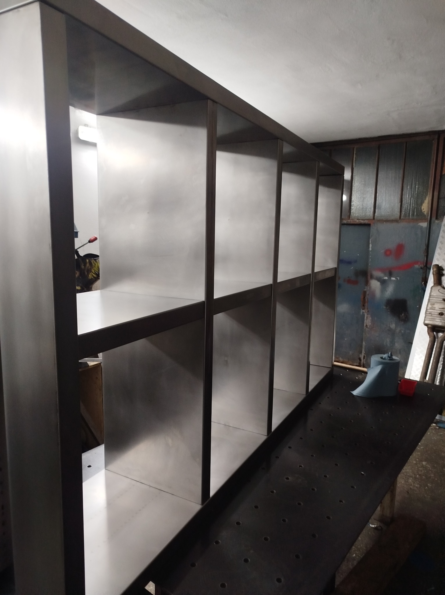 Metalowa konstrukcja regału z półkami w warsztacie, widoczne ślady obróbki i narzędzia w tle.