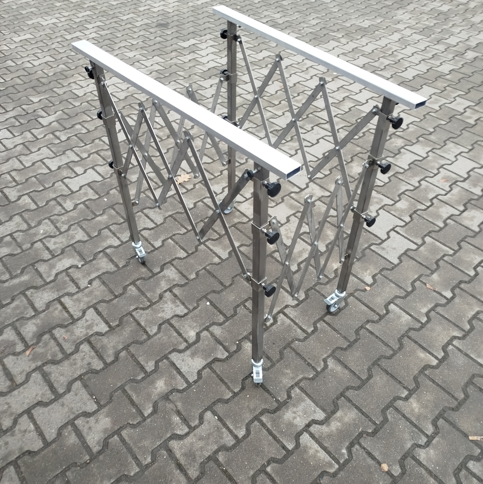 Rozkładana, mobilna konstrukcja z metalowych profili, na kółkach, z regulowanymi zaciskami, ustawiona na szarej kostce brukowej.