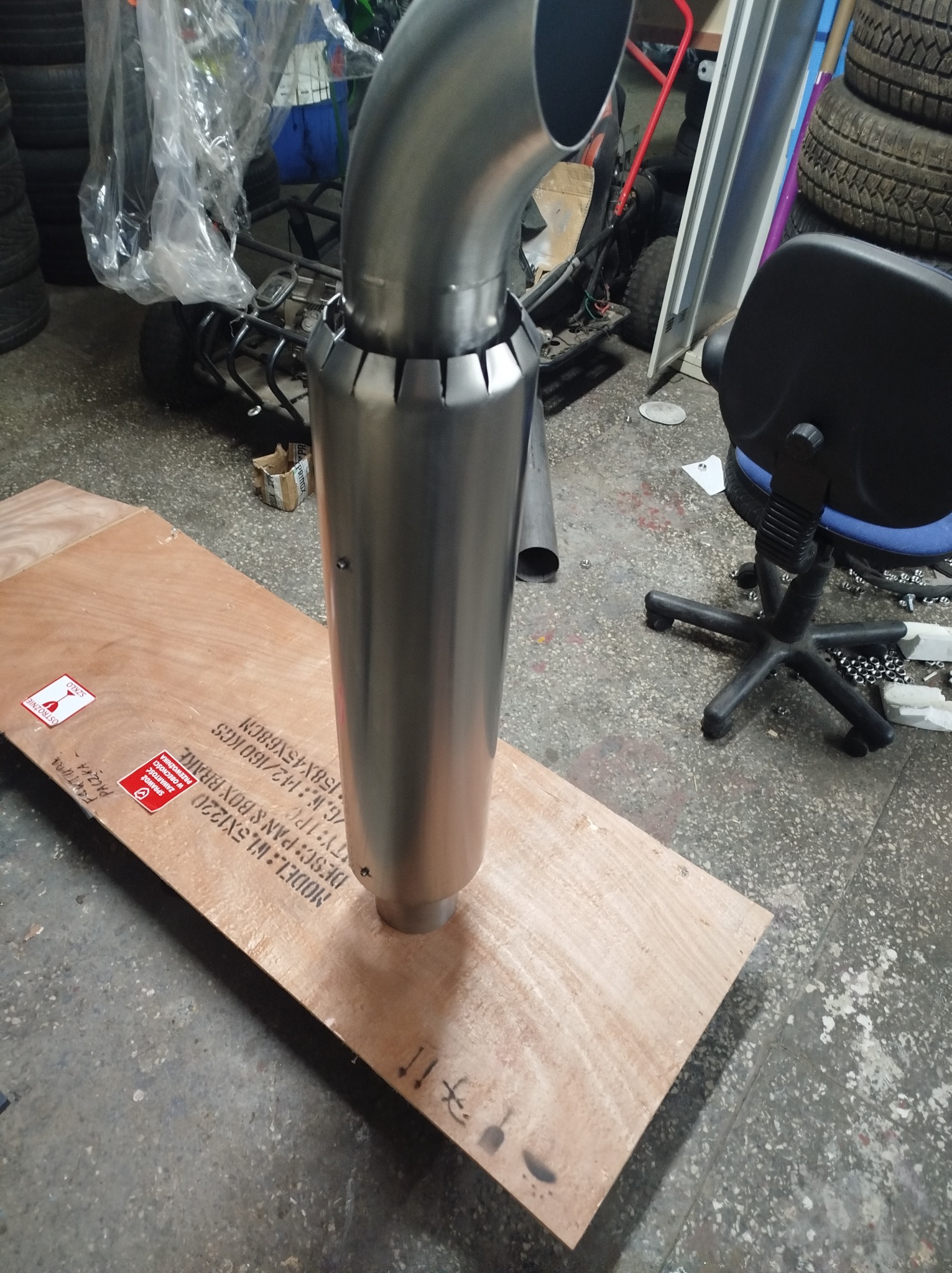 Nowy, błyszczący, metalowy element konstrukcyjny, prawdopodobnie część wydechu lub komina, stojący na drewnianej płycie w warsztacie, z widocznymi śladami użytkowania na podłodze i oponami w tle.