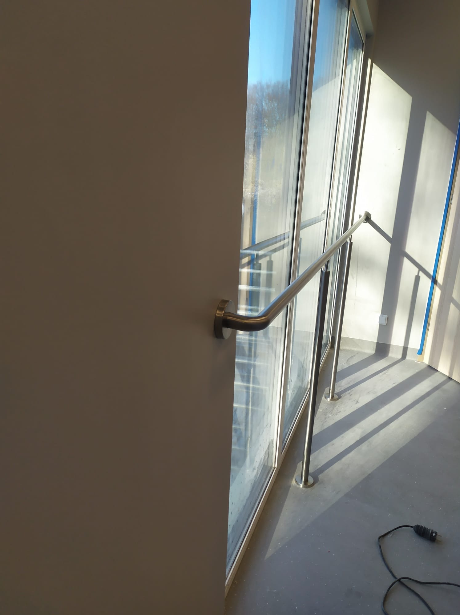 Wnętrze budynku z przeszkloną ścianą i zamontowaną metalową poręczą, widoczne światło słoneczne padające na podłogę i ścianę, kabel elektryczny leżący na podłodze.