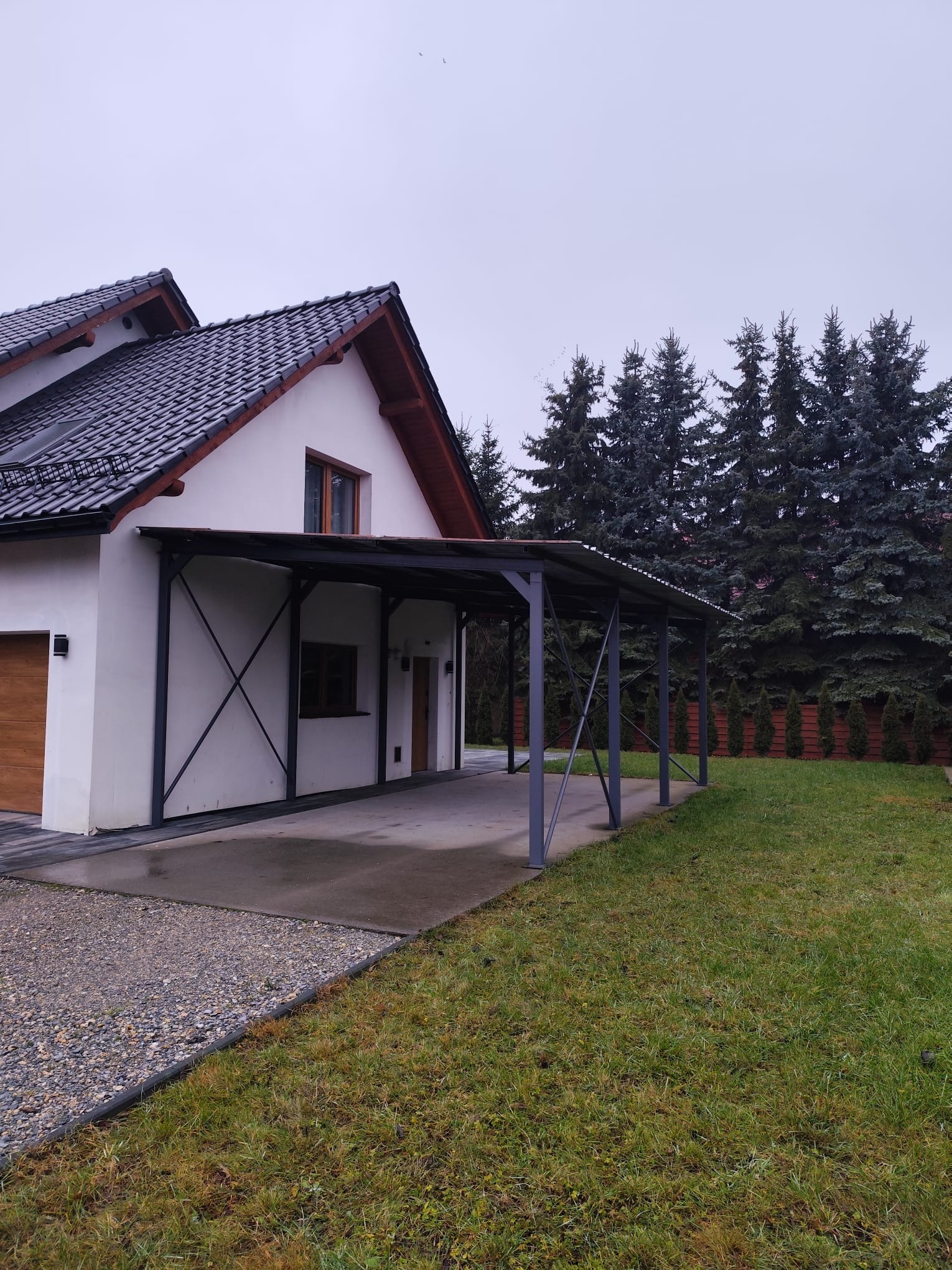 Nowoczesna, metalowa pergola przy białym domu z drewnianą bramą garażową. Szara konstrukcja z zadaszeniem, widoczne elementy mocujące. Tło: zielony trawnik i wysokie drzewa.