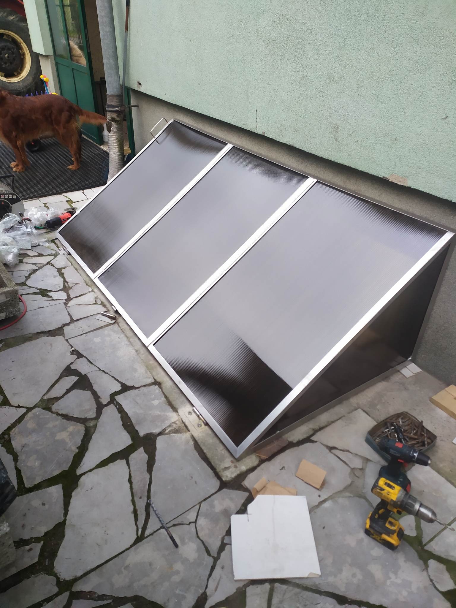 Zadaszenie zejścia do piwnicy z metalową ramą i wypełnieniem z tworzywa, zamontowane przy ścianie budynku. Na ziemi leżą narzędzia.