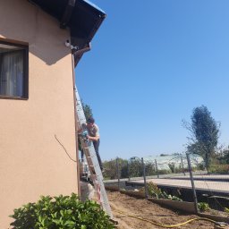 SECURE CAM - Pracownik na drabinie montuje kamerę monitoringu na elewacji domu, widoczne dwie zainstalowane kamery, na ziemi leży żółty kabel.