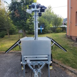 SECURE CAM - Mobilny system monitoringu wizyjnego na przyczepie z panelami słonecznymi, ustawiony na asfalcie przed budynkiem i zielenią.