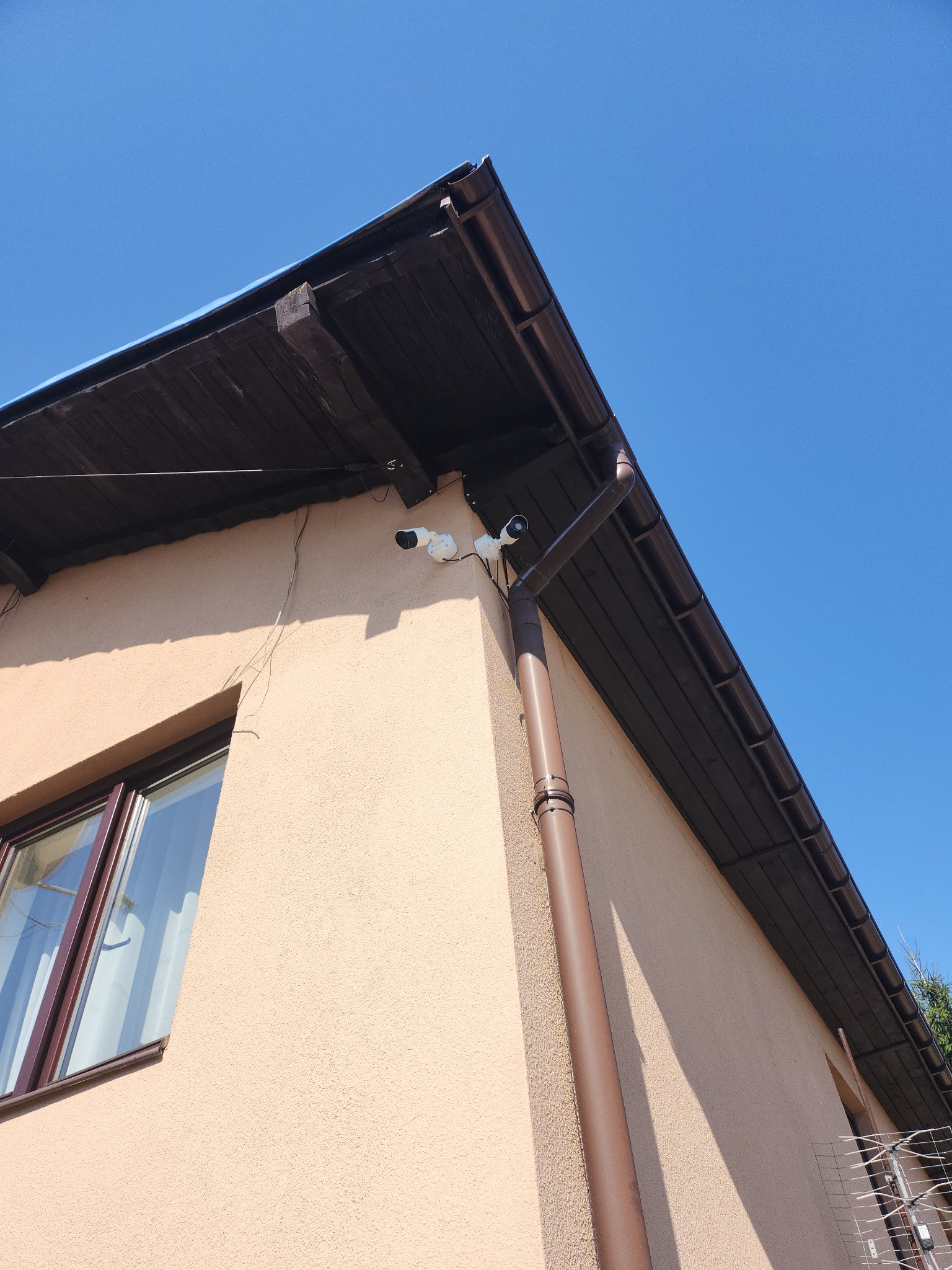 Dwie kamery monitoringu zamontowane na rogu domu z beżową elewacją, widoczne rynna i fragment dachu oraz antena telewizyjna w tle, na tle błękitnego nieba.