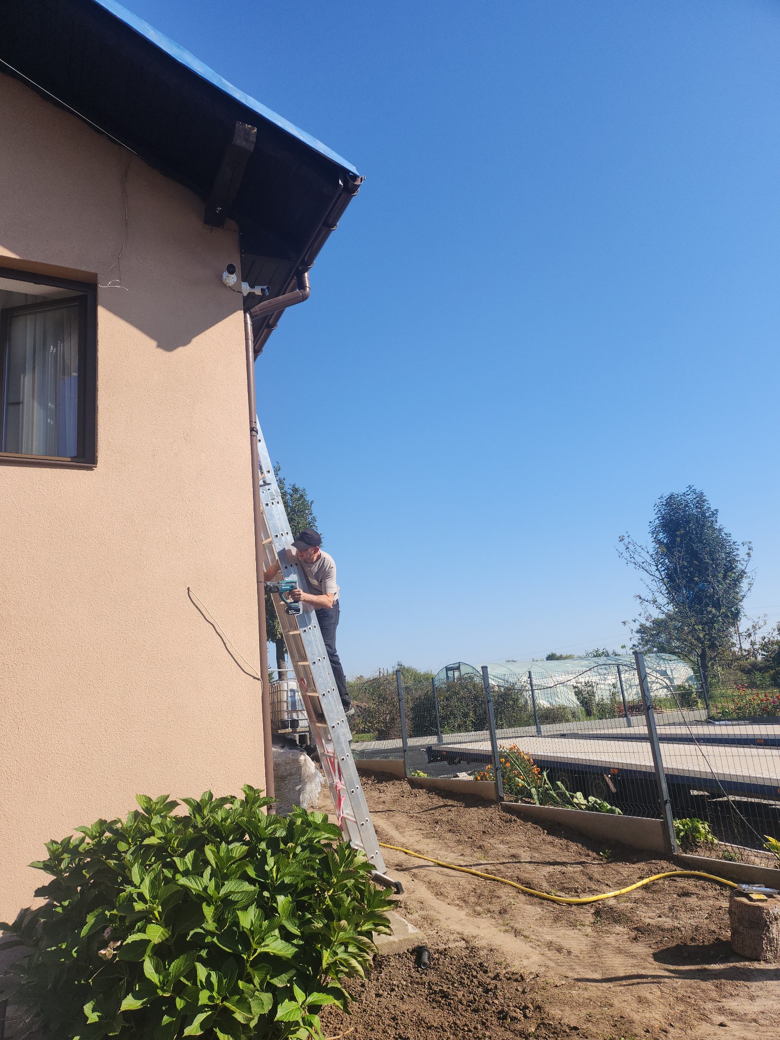 Pracownik na drabinie montuje kamerę monitoringu na elewacji domu, widoczne dwie zainstalowane kamery, na ziemi leży żółty kabel.