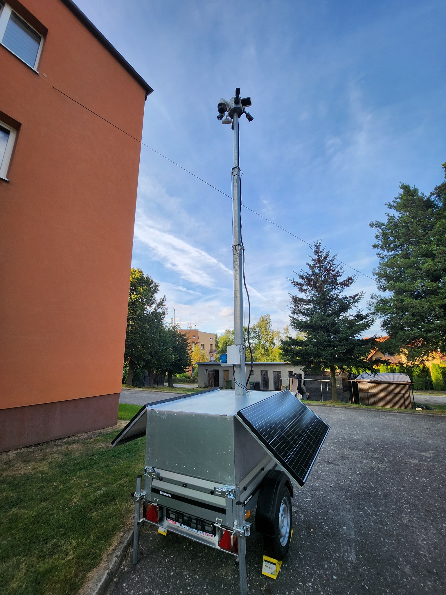 Mobilny system monitoringu wizyjnego na przyczepie z panelami słonecznymi, wysuwany maszt z kamerami, tło: budynek i drzewa.