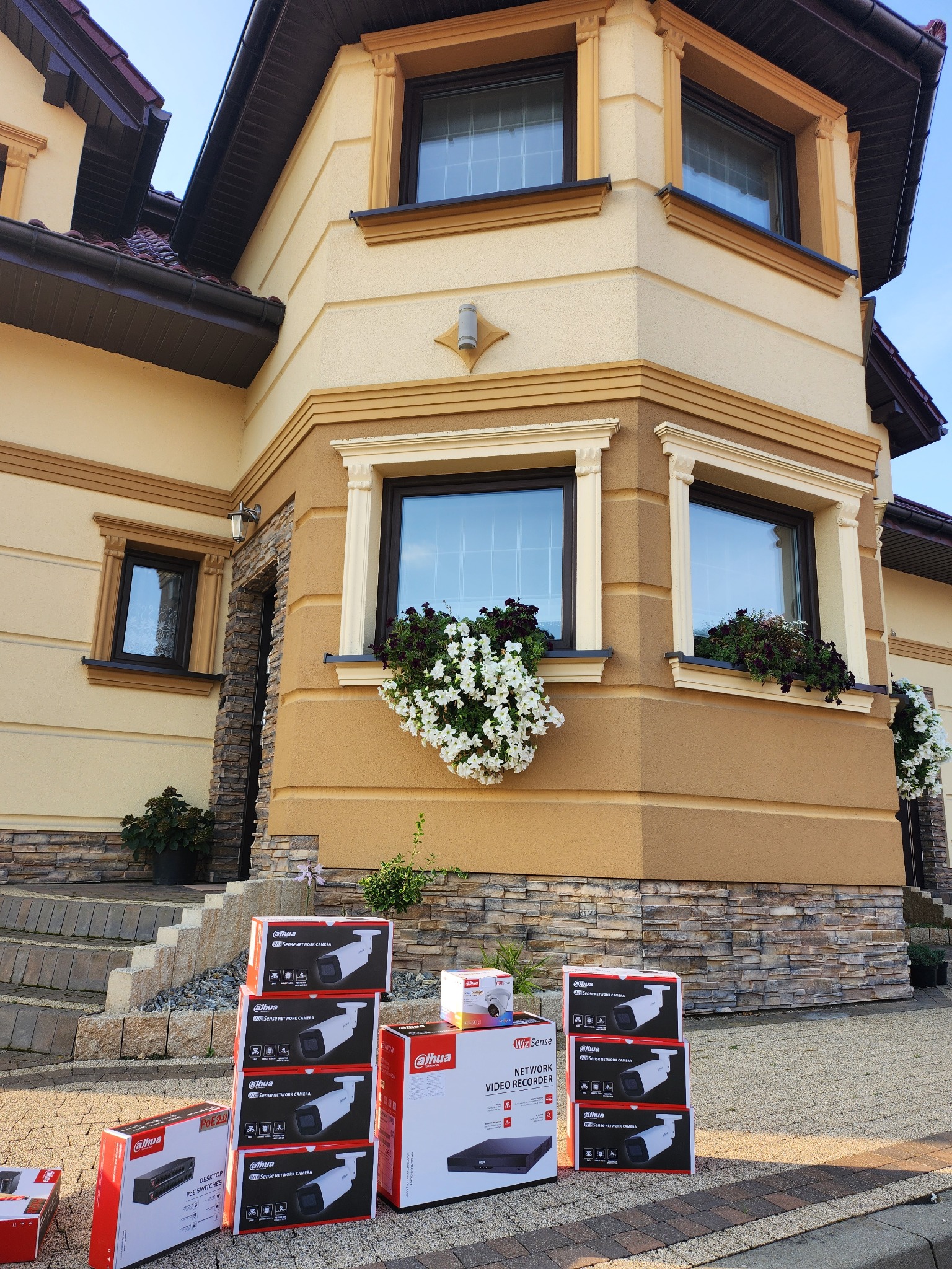 Pudełka z kamerami i rejestratorem firmy Dahua ustawione przed elewacją domu z kwiatami w oknach, sugerujące przygotowanie do instalacji systemu monitoringu.