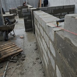 Usługi ogólnobudowlane Szymon Płotka - Wznoszenie ściany z szarych bloczków betonowych, widoczna rozciągnięta czerwona linka murarska, w tle betonowa ściana i pracownik na rusztowaniu.