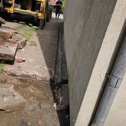 Usługi ogólnobudowlane Szymon Płotka - Żółta koparka Volvo z operatorem w kabinie, przygotowująca teren obok budynku z rurą spustową i wykopanym rowem, z widocznymi cegłami i mężczyzną w tle.