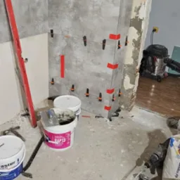 Szare płytki ścienne układane z użyciem poziomicy, krzyżyków dystansowych i taśmy, widoczne wiaderka z zaprawą, mieszadło i inne narzędzia na podłodze w trakcie prac wykończeniowych.