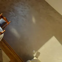 Beżowa ściana z dekoracyjną strukturą, fragment drewnianej podłogi i narożnika sofy z obrazami w złotych ramach, widok z góry.