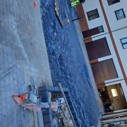 Usługi ogólnobudowlane Szymon Płotka - Układanie kostki brukowej na podjeździe, widoczny fragment ułożonej nawierzchni, maszyna do cięcia kostki, paleta z kostką, pracownik w odblaskowej kamizelce z daleka.