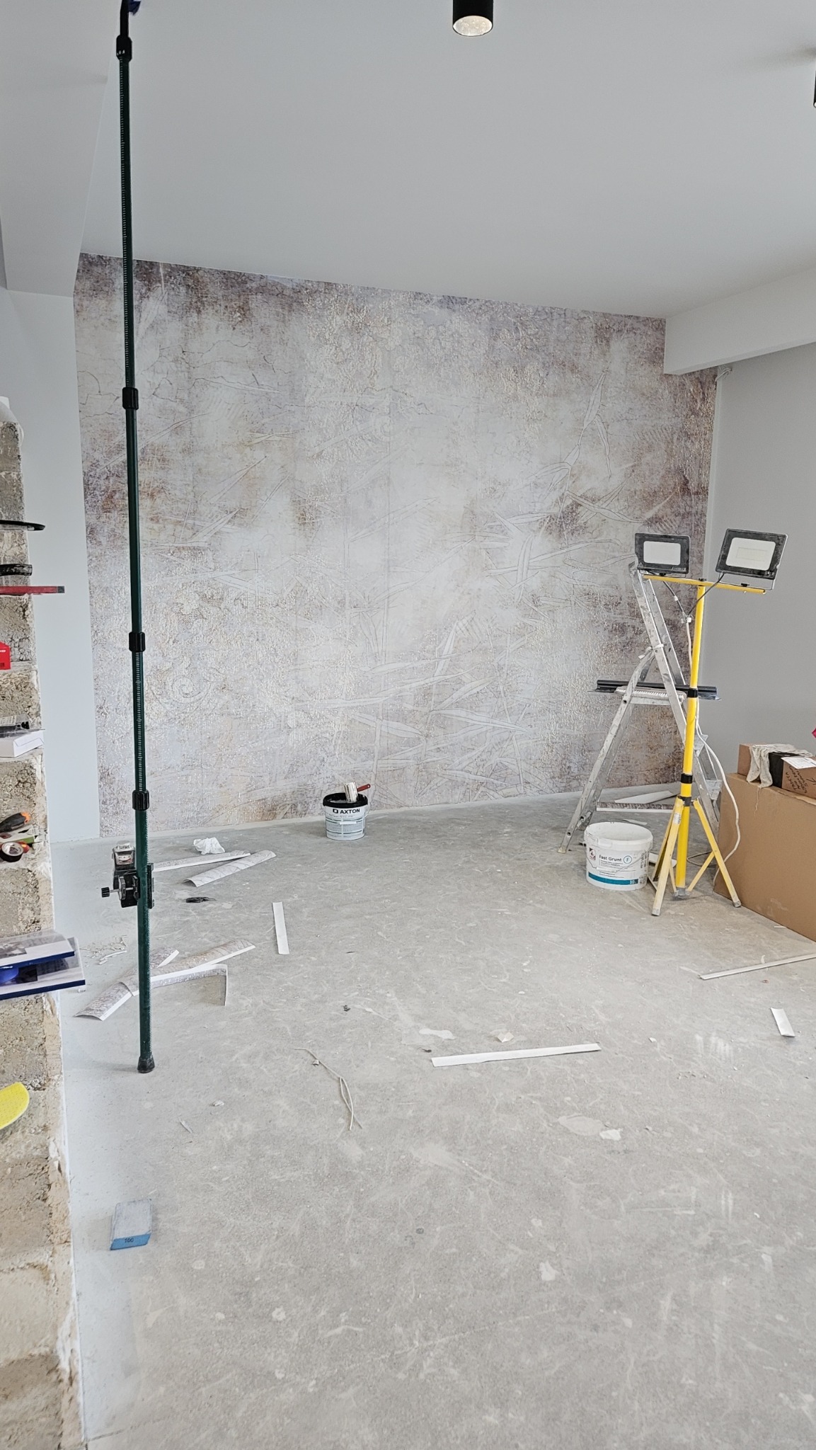 Pokój w trakcie prac wykończeniowych: ściana z dekoracyjną tapetą, drabina z lampami roboczymi, puszki z farbą Axton i gruntem, listwy przypodłogowe na podłodze, widoczny teleskopowy kij malarski.