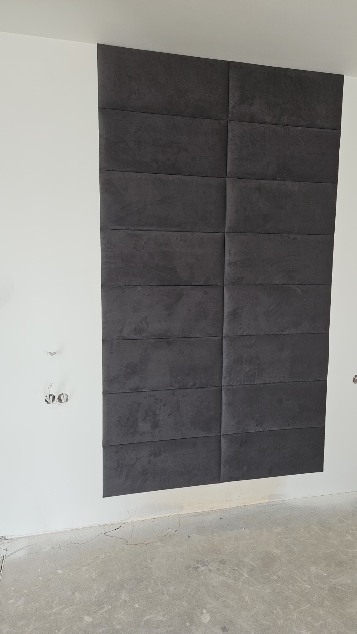 Ściana z zamontowanymi, ciemnoszarymi panelami tapicerowanymi w układzie prostokątnym, widoczne puszki elektryczne bez osprzętu i surowa podłoga.