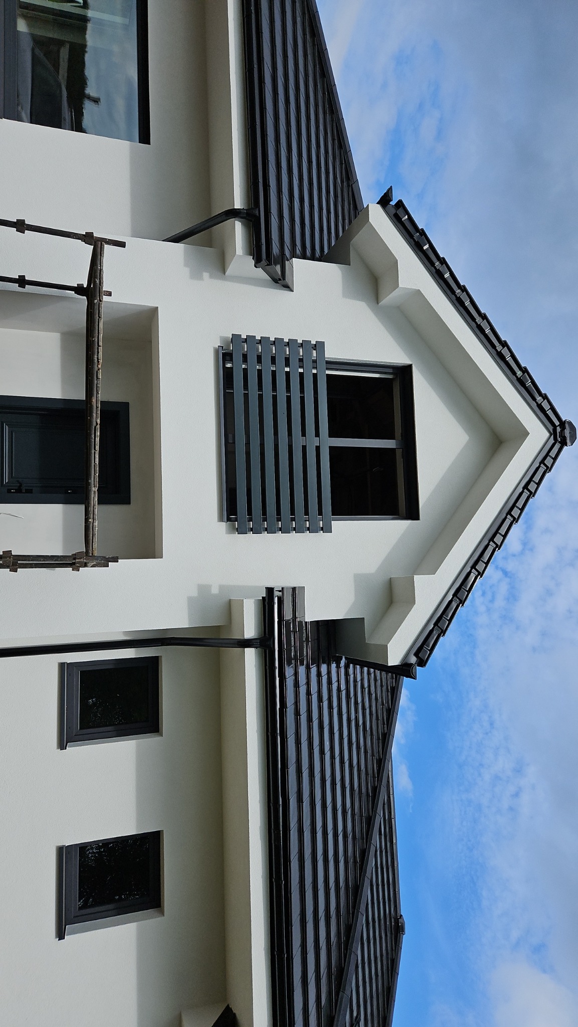 Biała fasada domu z ciemnymi oknami, żaluzjami i dachem, widoczne rusztowanie z boku.