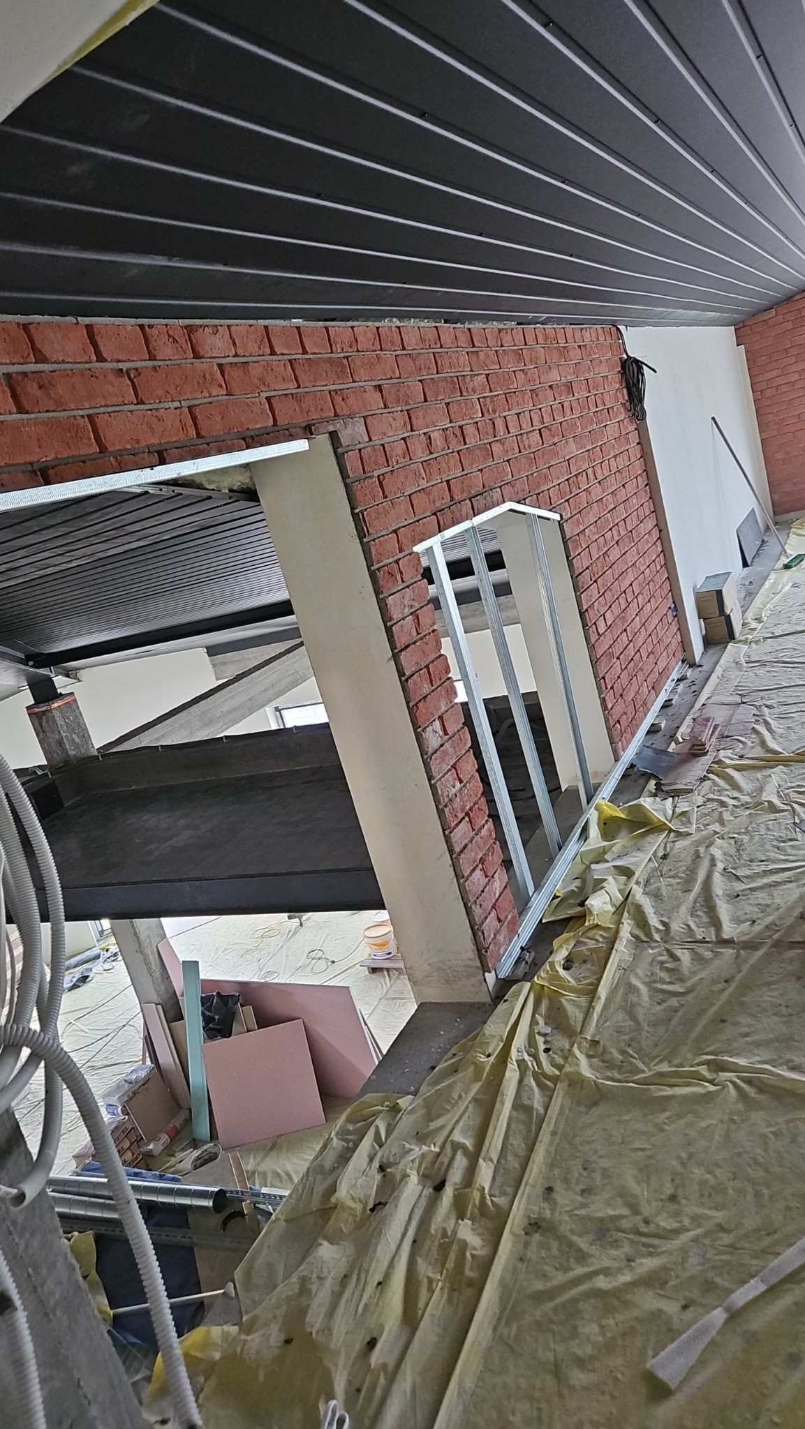 Wnętrze w trakcie prac wykończeniowych: murowana ściana z czerwonej cegły, metalowa konstrukcja okienna, zabezpieczona folią podłoga, widoczne materiały budowlane i czarne panele sufitowe.