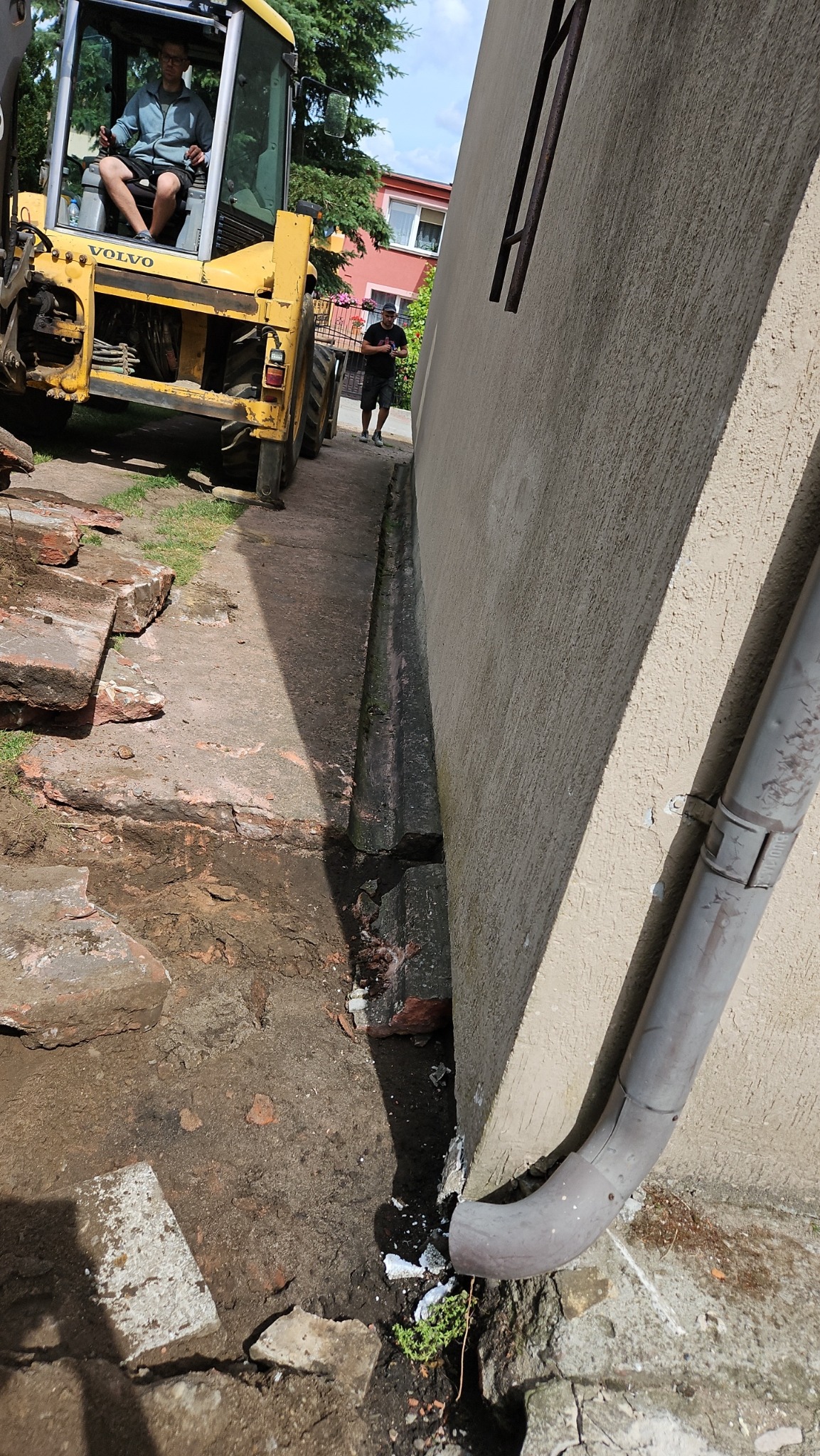 Żółta koparka Volvo z operatorem w kabinie, przygotowująca teren obok budynku z rurą spustową i wykopanym rowem, z widocznymi cegłami i mężczyzną w tle.