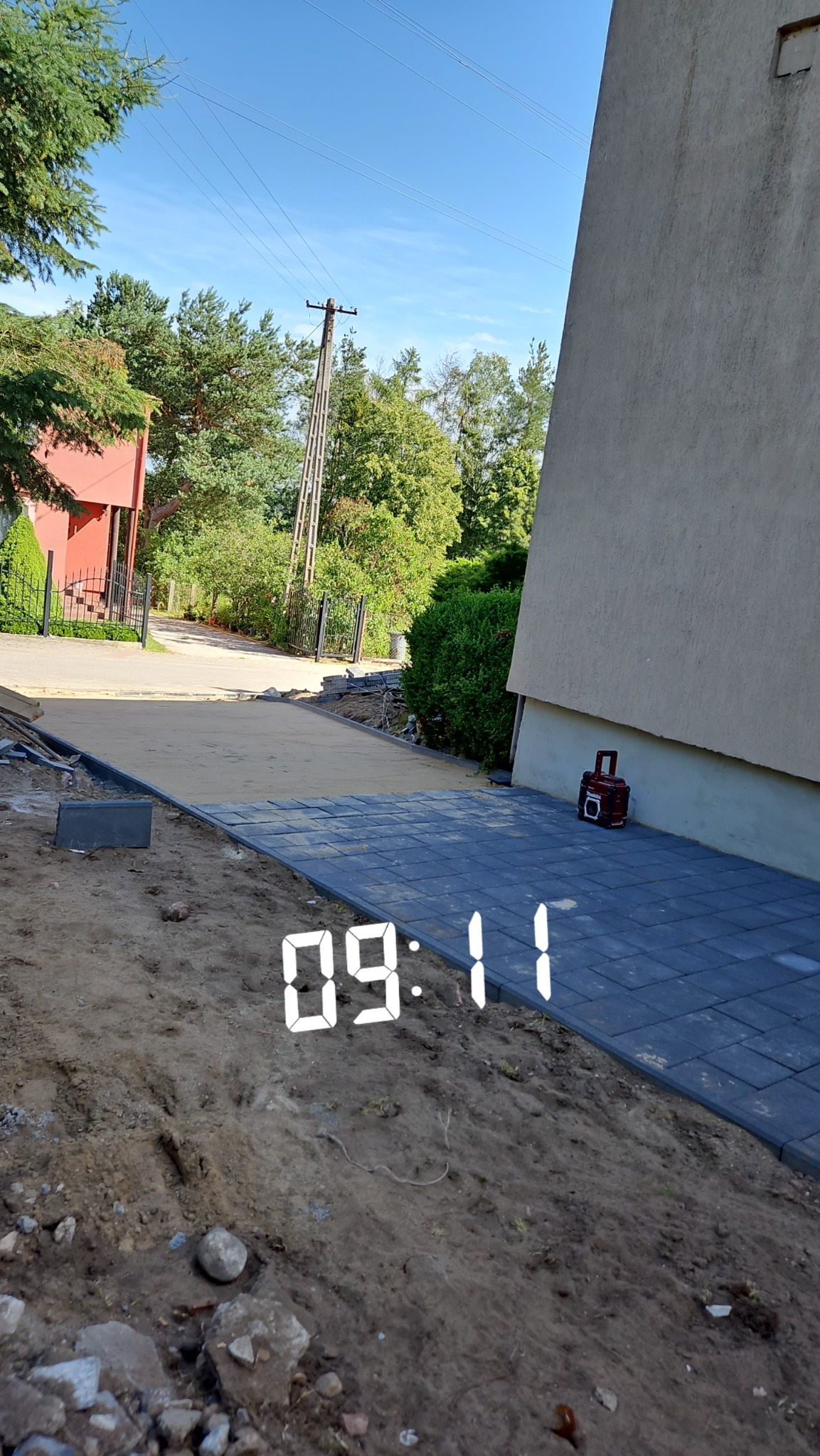 Układanie szarej kostki brukowej na podwórku w trakcie realizacji, widoczny fragment budynku i piasek pod kostką, w tle skrzynka narzędziowa.