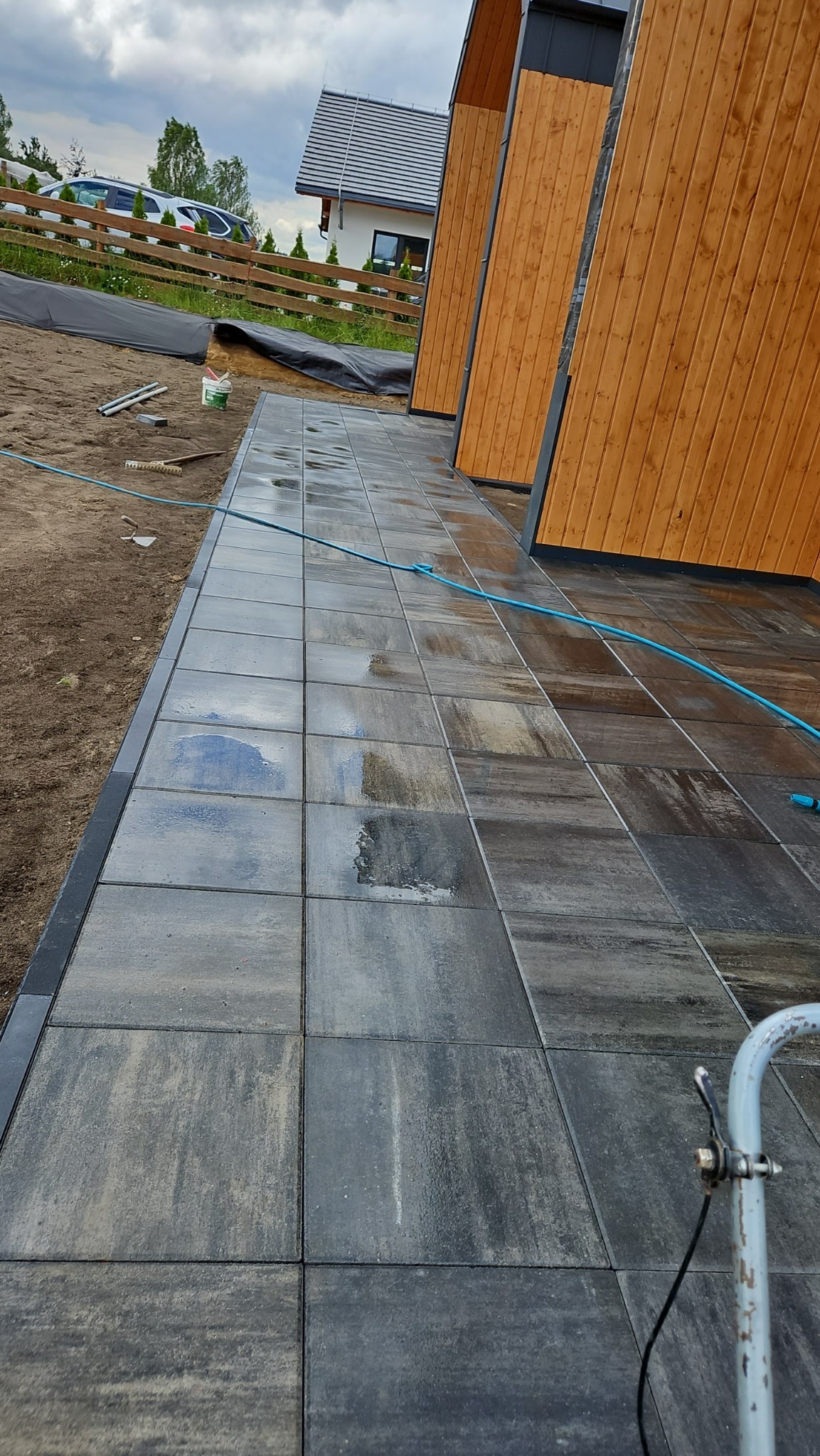 Ułożona, mokra nawierzchnia z szarych płyt betonowych przy drewnianej elewacji budynku. Widoczny wąż ogrodowy, ziemia i fragment domu w tle.