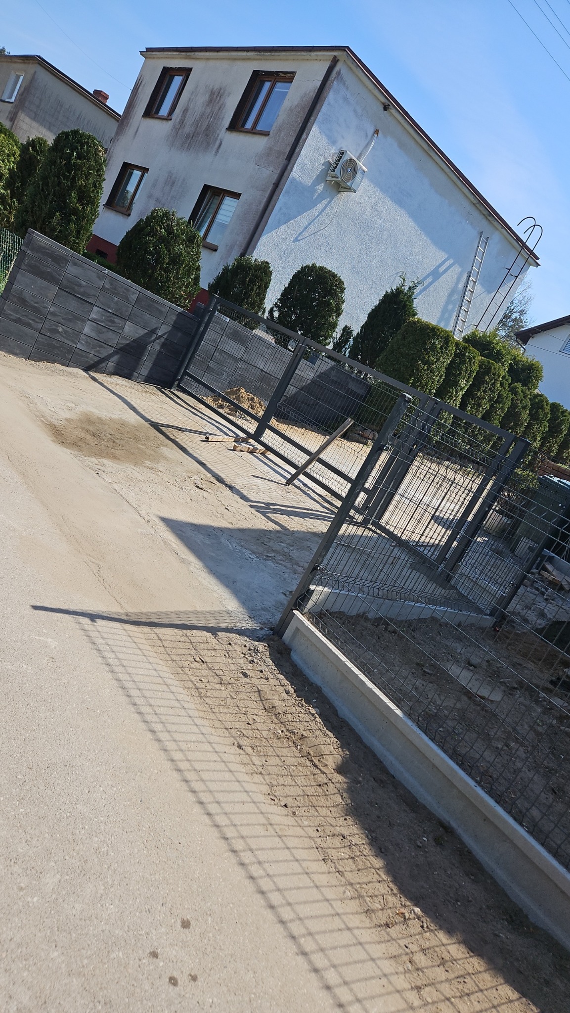 Montaż metalowego ogrodzenia panelowego na betonowej podmurówce, widok na dom z klimatyzacją i tujami w tle.