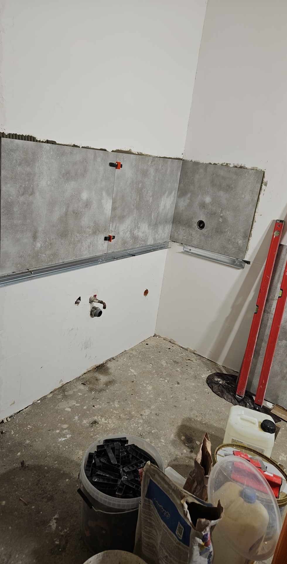 Fragment ściany w trakcie układania szarych płytek z użyciem poziomicy i systemu poziomowania; widoczne puszki instalacyjne i wiadro z klinami do płytek.