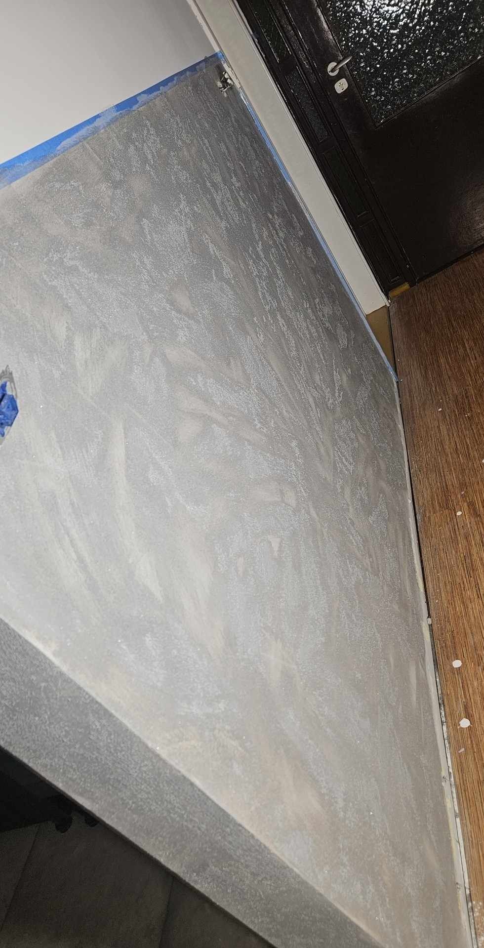 Ściana pokryta dekoracyjną, jasnoszarą gładzią z efektem delikatnego wzoru, zabezpieczona niebieską taśmą malarską przy suficie, widoczny fragment drewnianej podłogi i czarne drzwi.