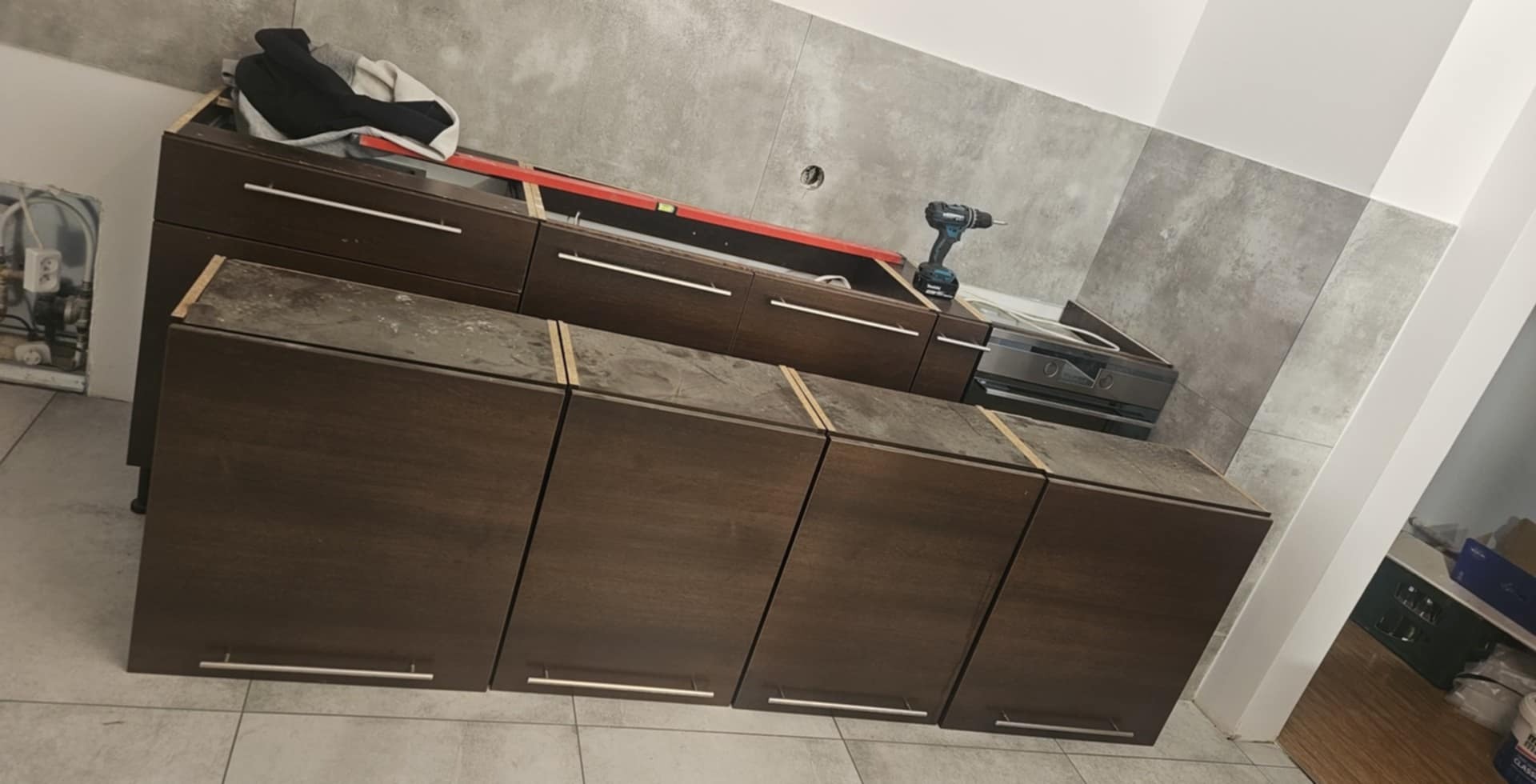 Montaż brązowych szafek kuchennych z poziomnicą i wiertarką na blacie, szare płytki na ścianie w tle.