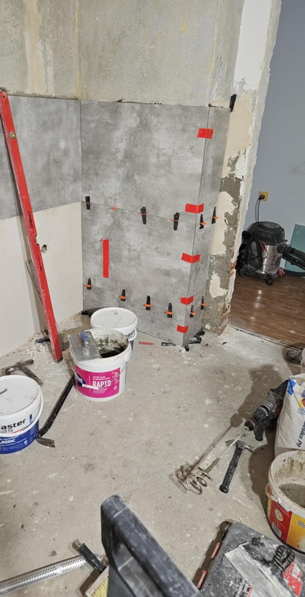 Szare płytki ścienne układane z użyciem poziomicy, krzyżyków dystansowych i taśmy, widoczne wiaderka z zaprawą, mieszadło i inne narzędzia na podłodze w trakcie prac wykończeniowych.