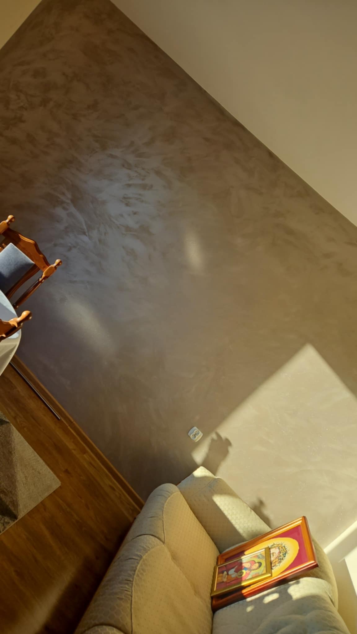 Beżowa ściana z dekoracyjną strukturą, fragment drewnianej podłogi i narożnika sofy z obrazami w złotych ramach, widok z góry.