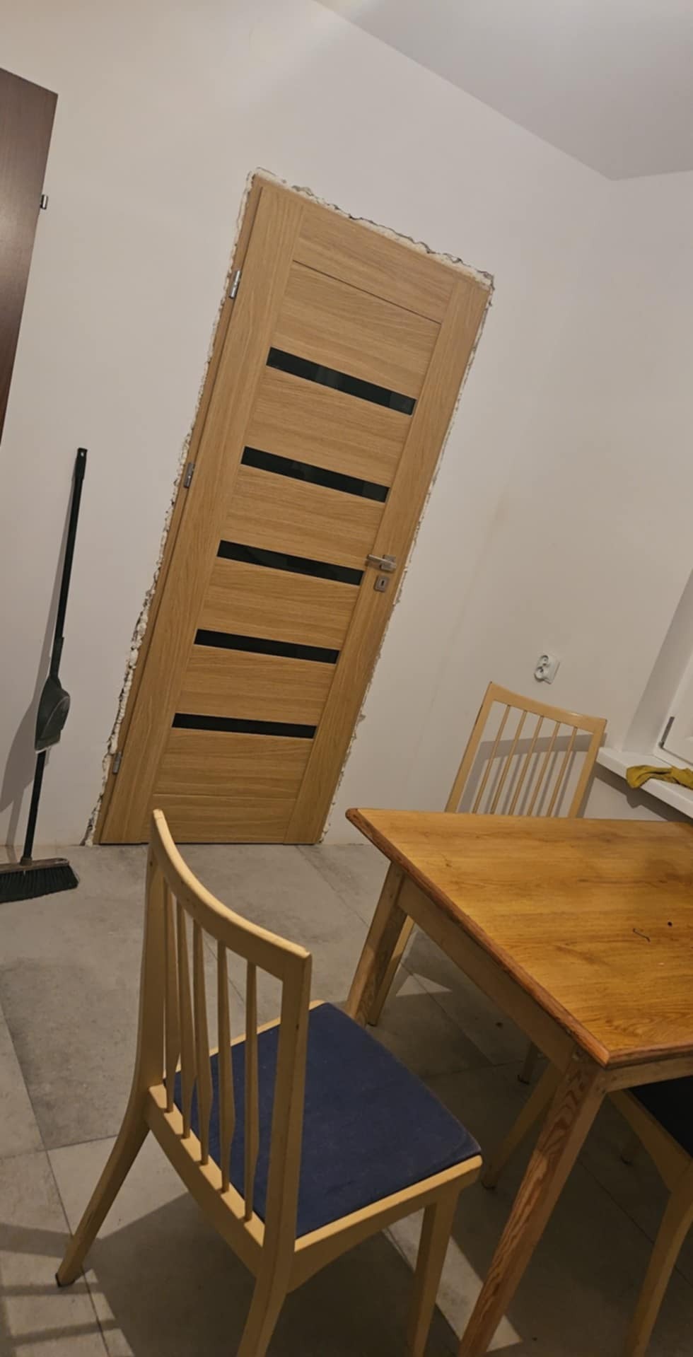 Montaż ościeżnicy drzwiowej z widocznymi szczelinami i resztkami pianki montażowej, w tle drewniany stół i krzesła, pod ścianą stoi zmiotka.